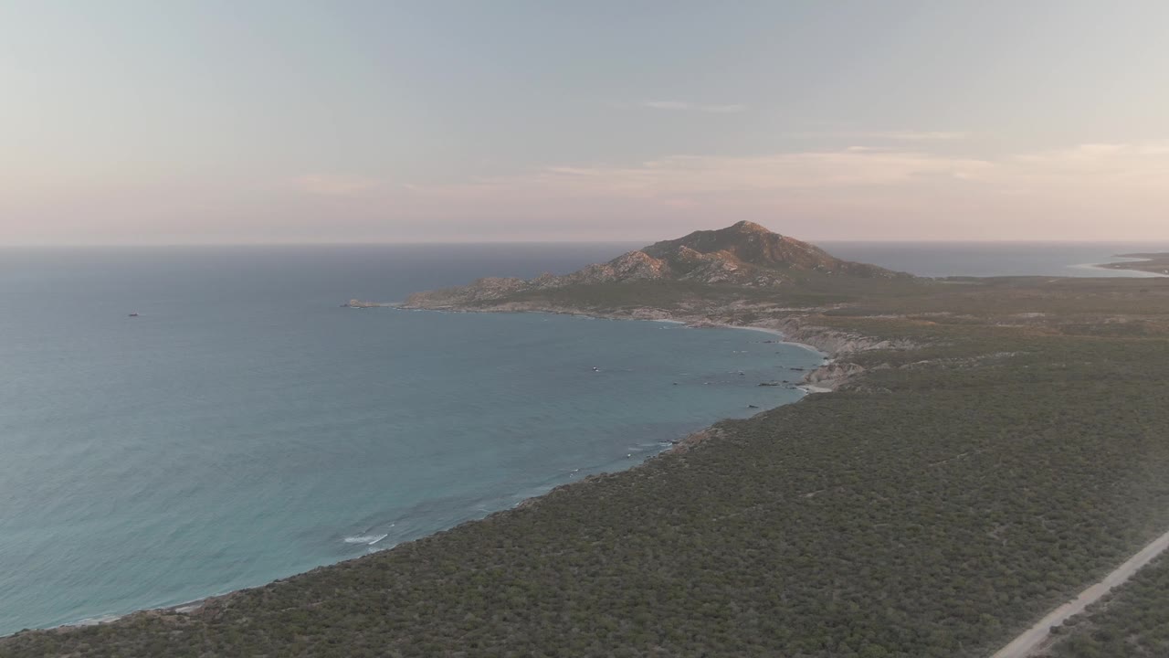 High aerial frame over Cabo Pulmo with blue ocean filling the left side, scrub-covered land and a pale road in the lower right, a beach arc at midframe, a rocky hill beyond, and no visible people.