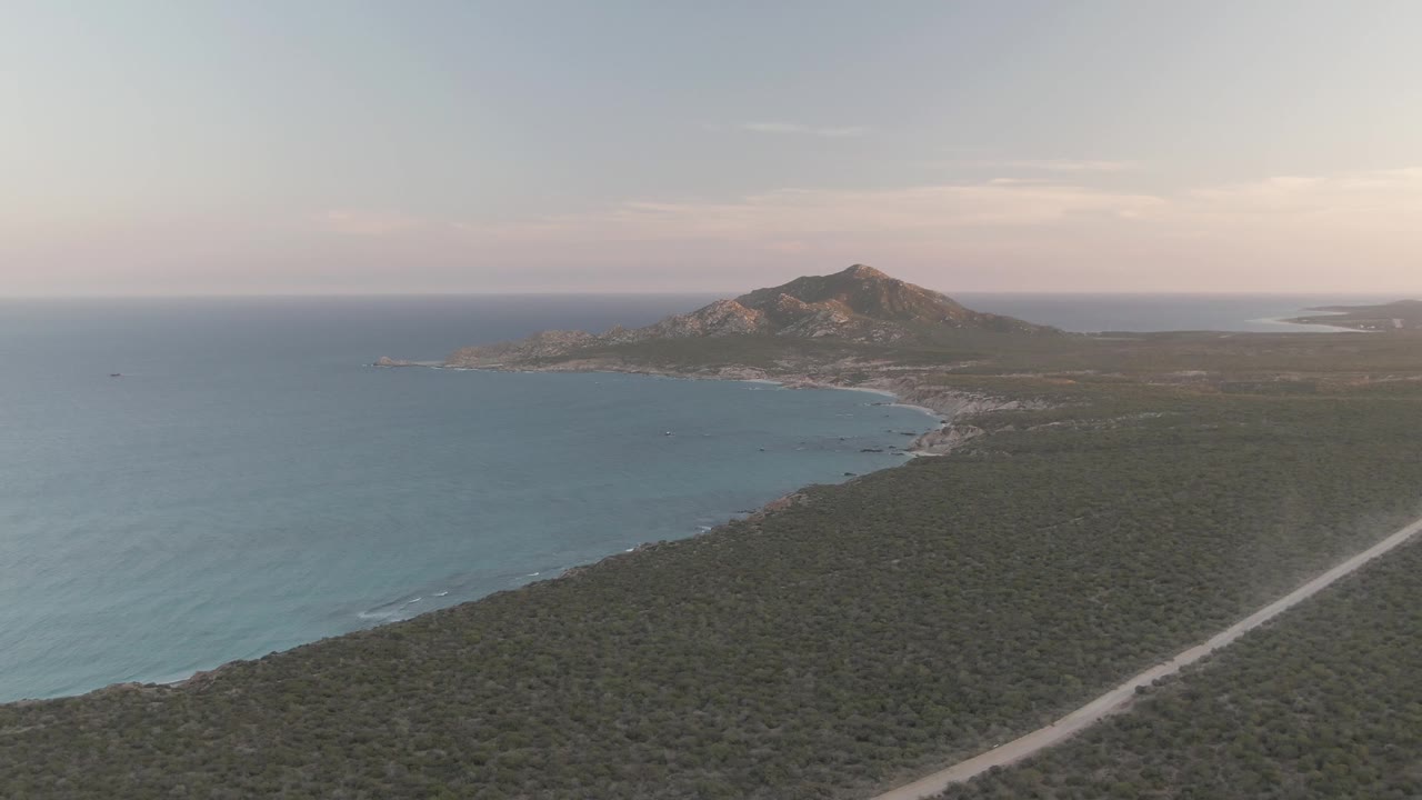Aerial view in Cabo Pulmo, Baja California Sur with blue ocean on the left, a pale road cutting through green scrub, a curved beach, a rocky hill in the distance, and no visible people.