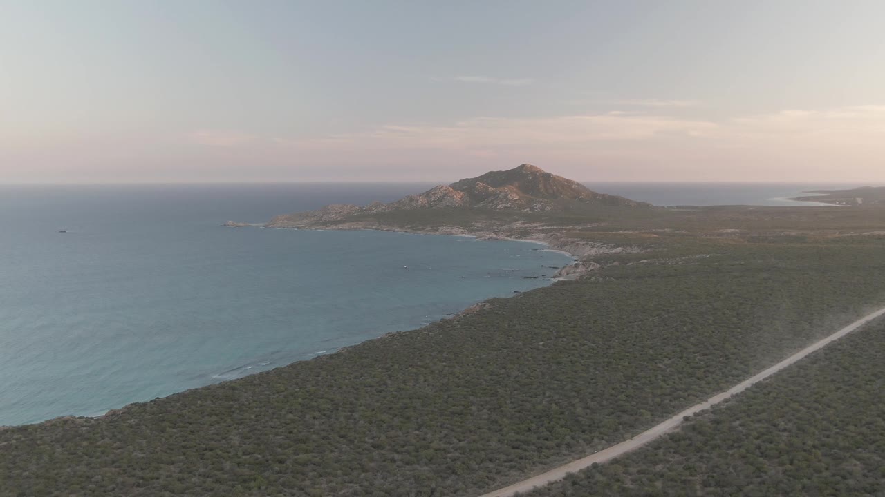 Cabo Pulmo aerial frame with the coastline curving beneath a rocky hill, open blue water to the left, scrubland across the foreground, a pale road along the right side, and no visible people.
