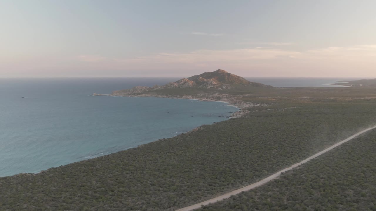 High oblique Cabo Pulmo view with smooth blue water, a narrow beach curve, dense green scrub, the pale road tracking diagonally through the right foreground, and no visible people.