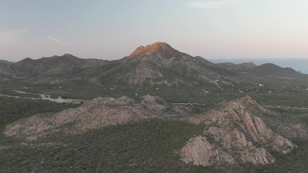 Wide aerial view in Cabo Pulmo, Baja California Sur, with rocky green hills in the foreground, a warm-lit mountain peak beyond the valley, faint sea on the horizon, and no visible people.