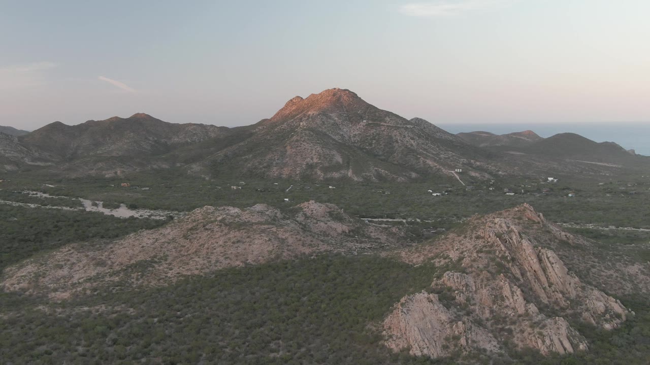 Aerial Cabo Pulmo landscape with a rocky ridge lower in frame, dense green valley cover, a pale riverbed crossing left of center, and a dusk-lit mountain peak with no visible people.