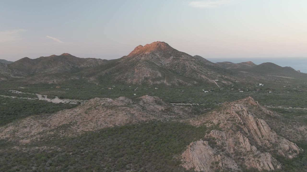 Cabo Pulmo aerial frame showing scrub-covered hills, exposed rock faces, a central mountain peak touched by warm dusk light, pale sky, distant water, and no visible people.