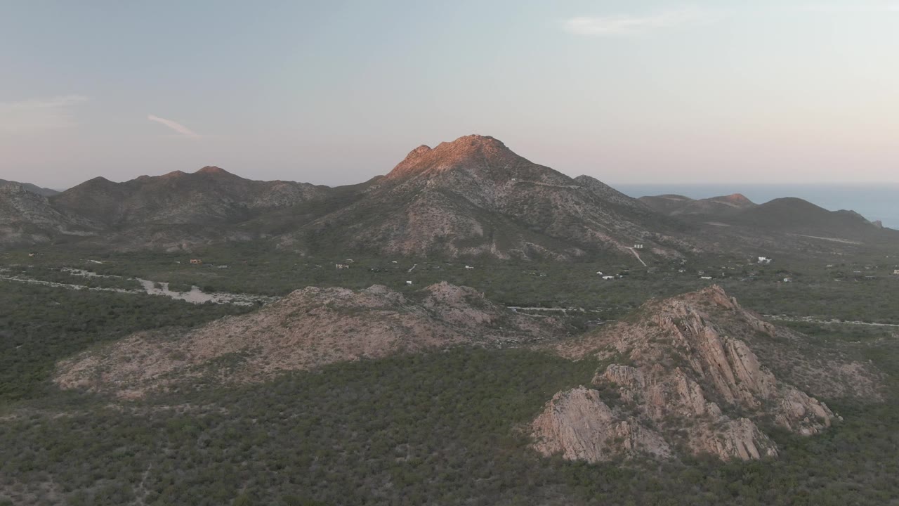 Aerial frame over Cabo Pulmo showing rocky foreground ridges, scrub-covered valley terrain, a pale road, distant mountains, soft evening sky, and no visible people.