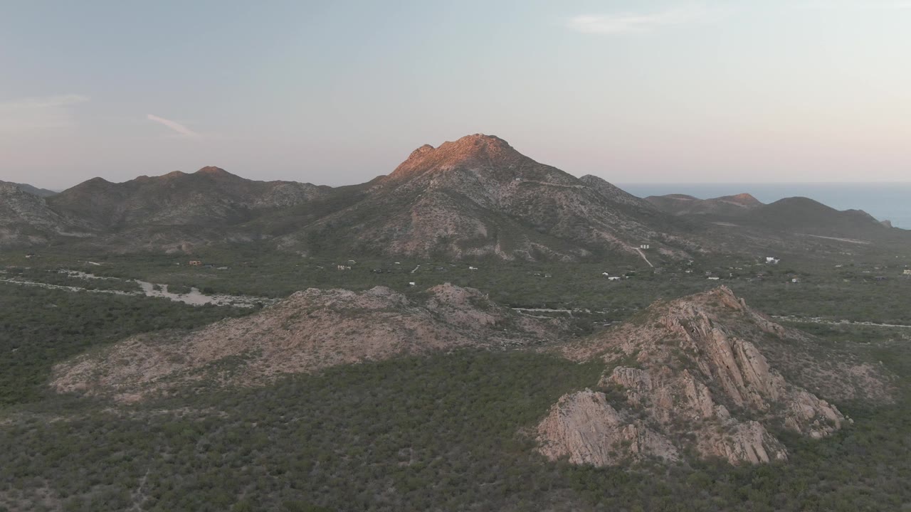 Aerial view in Cabo Pulmo with the warm-lit mountain peak centered higher in frame, rocky ridges below, a road through the valley, and no visible people.