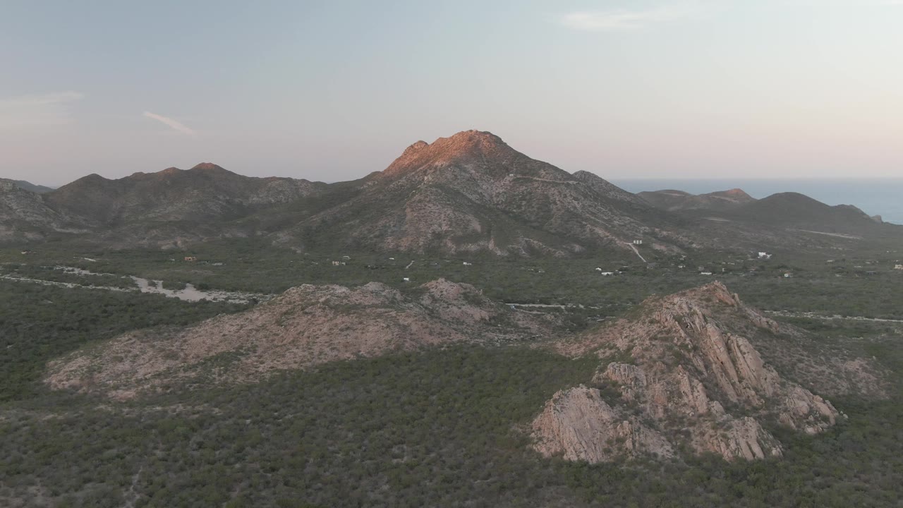 Cabo Pulmo aerial frame showing the main mountain slightly right of center, the valley road with tiny vehicles, muted green vegetation, rocky slopes, and no visible people.