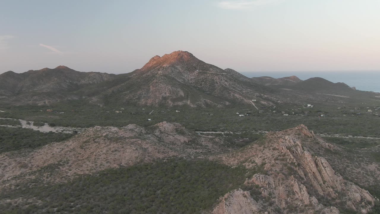 Opening frame over Cabo Pulmo shows rocky foreground ridges, a green valley road, a sunlit mountain peak, distant ocean horizon, and no visible people.