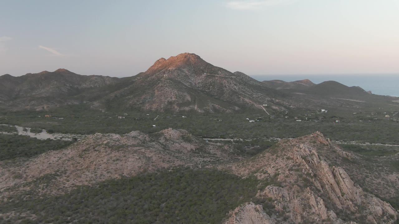 Cabo Pulmo aerial frame at one second centers the warm-lit peak above the valley road, with rocky ridges below, ocean in the distance, and no visible people.