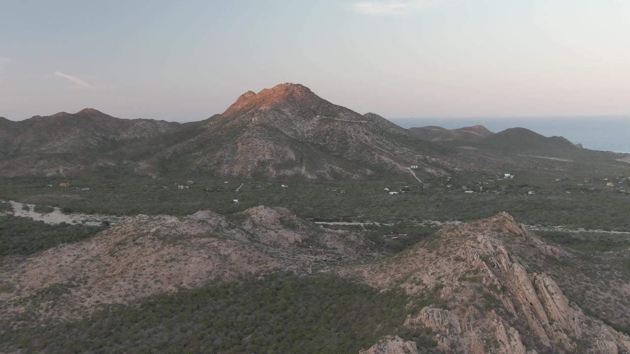 Aerial mountain landscape with rocky slopes, green valley floor, faint roads, and a soft sky.