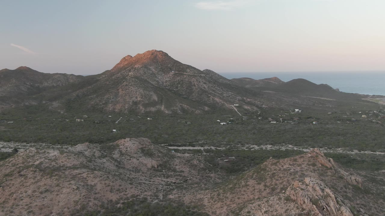 Early frame of Cabo Pulmo, Baja California Sur, with the rocky foreground ridge lower in frame, scattered valley structures, distant water, and no visible people.