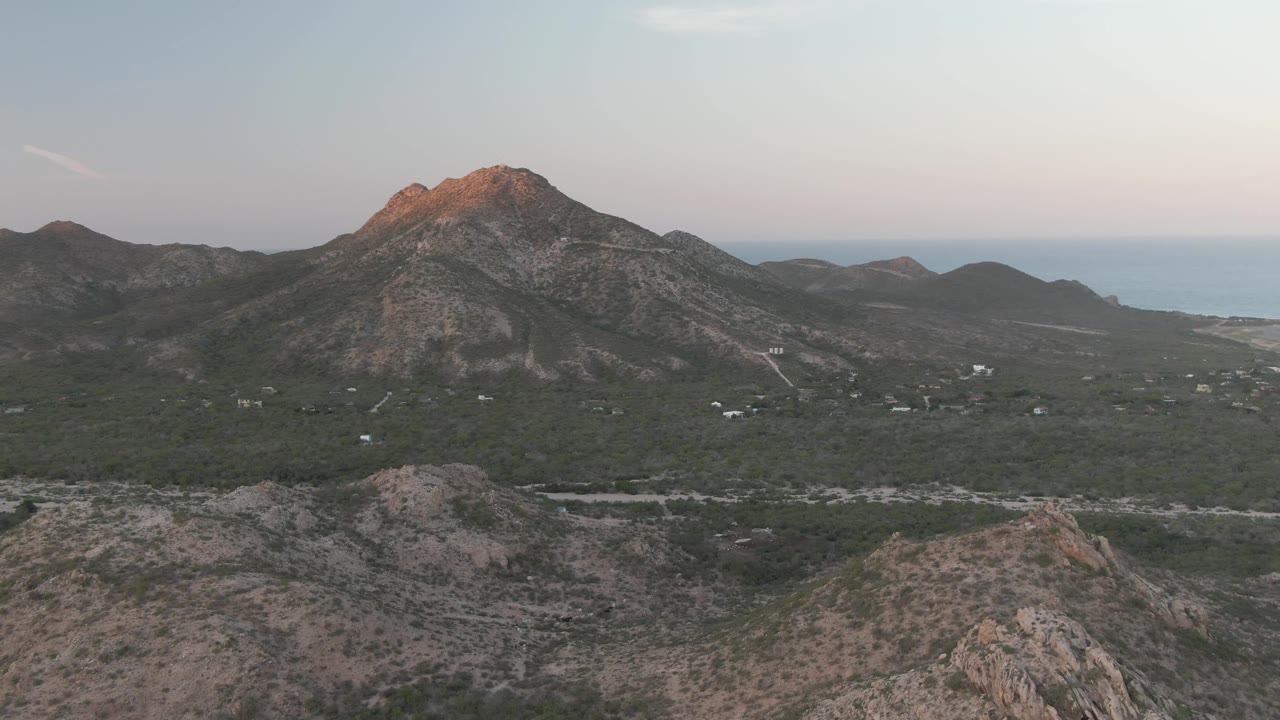 Cabo Pulmo mountain valley frame with layered rocky hills in front, a broad green midground, distant blue water on the right, and no visible people.
