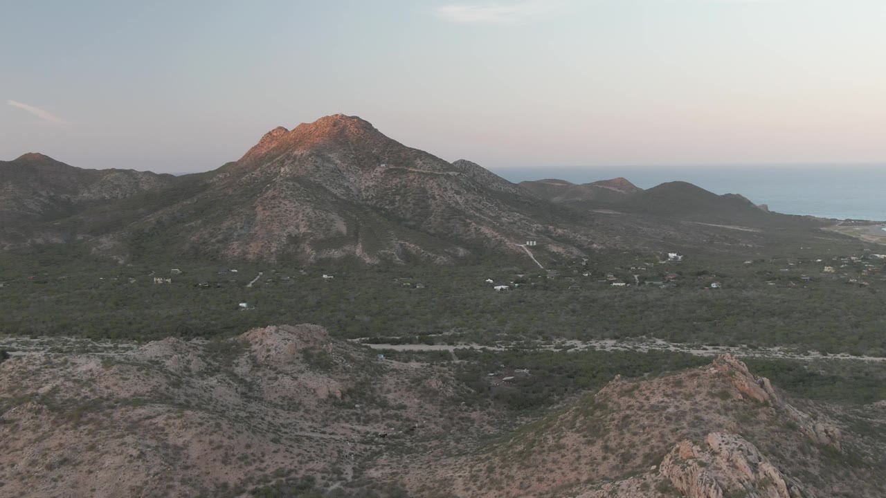 Mid-clip frame over Cabo Pulmo, Baja California Sur, showing the warm top of the mountain, dark green valley vegetation, rocky foreground terrain, and no visible people.
