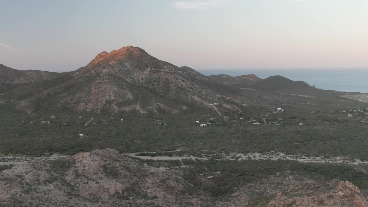 Opening aerial frame over Cabo Pulmo, Baja California Sur, with a rocky mountain centered beyond green scrubland, pale roads, distant water, and no visible people.