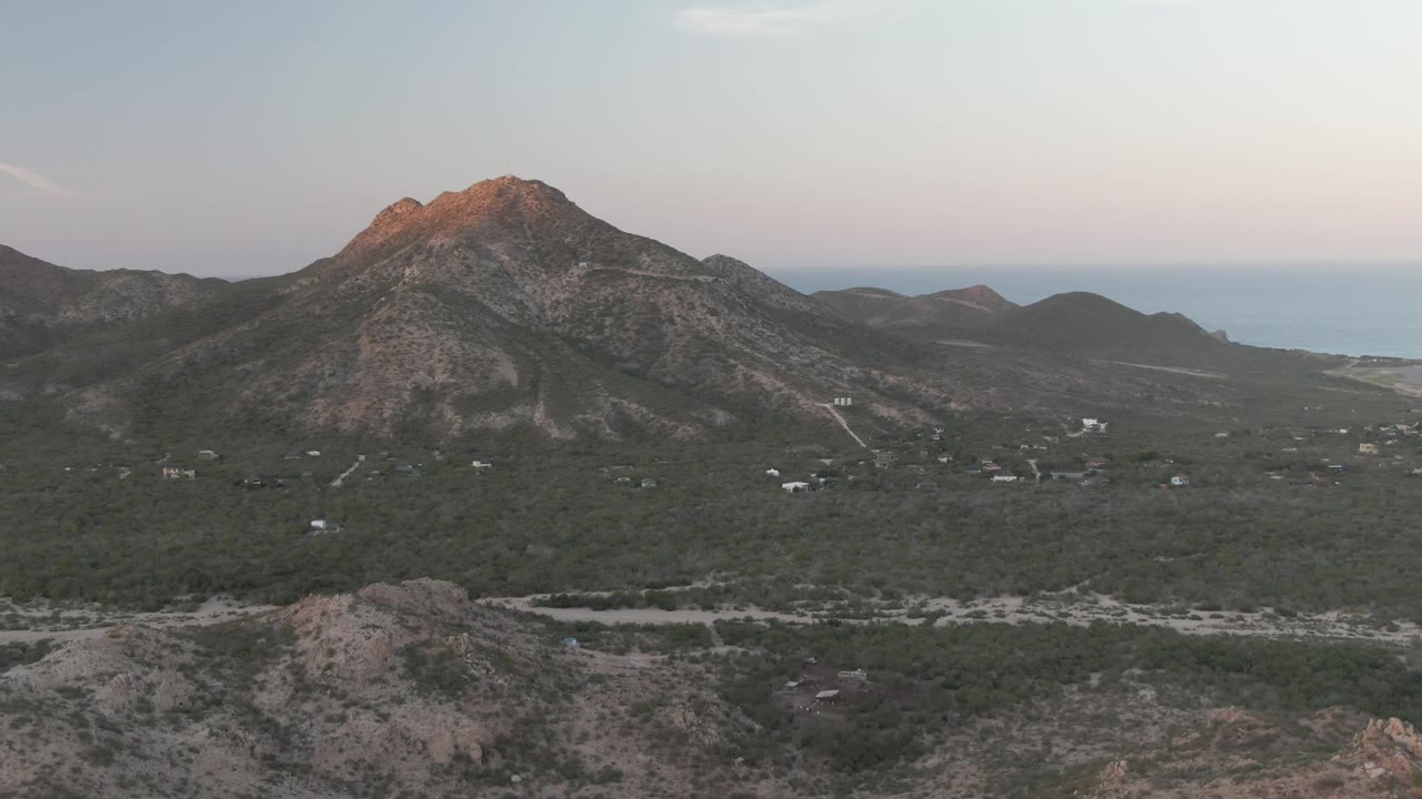 Early descent frame in Cabo Pulmo showing the central rocky ridge above dense green vegetation, a winding road, small distant buildings, and no visible people.