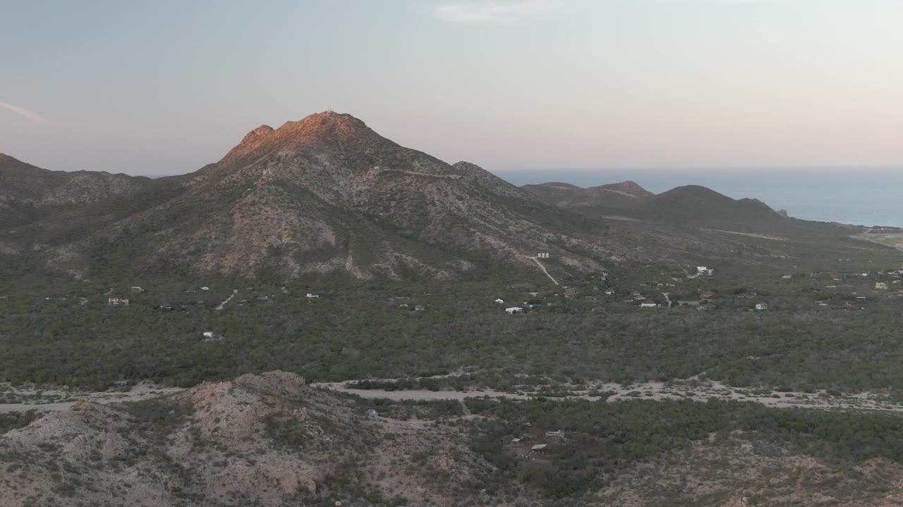 Mid-early frame over Cabo Pulmo with the camera looking across scrub-covered terrain toward a rocky mountain, distant coastline, pale roads, and no visible people.
