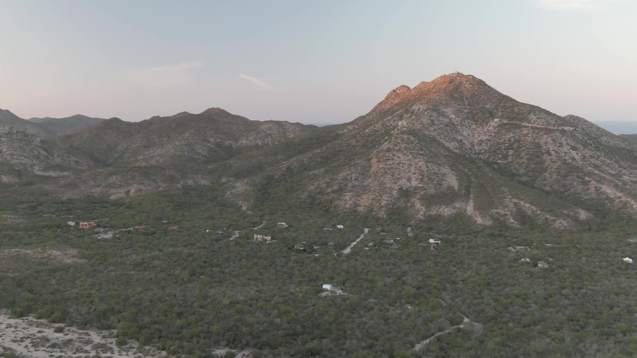 Aerial frame over Cabo Pulmo showing a green valley, scattered roads, distant hills, and a rocky peak lit by warm sunset color, with no visible people.