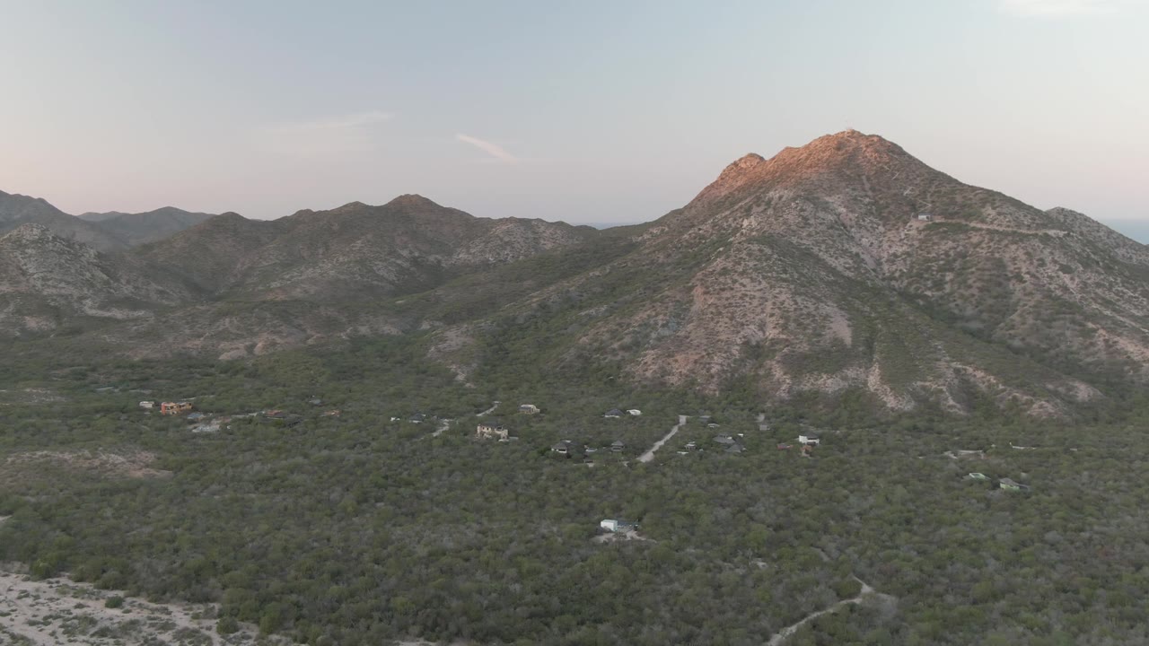 Wide aerial view in Cabo Pulmo with dense green vegetation below, pale roads crossing the valley, layered hills in the distance, and no visible people.