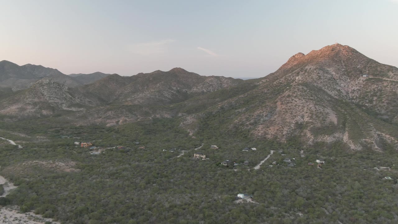 Opening aerial frame over Cabo Pulmo, Baja California Sur, showing dense green vegetation, scattered rural buildings, pale dirt roads, and rocky mountains under soft light, with no visible people.