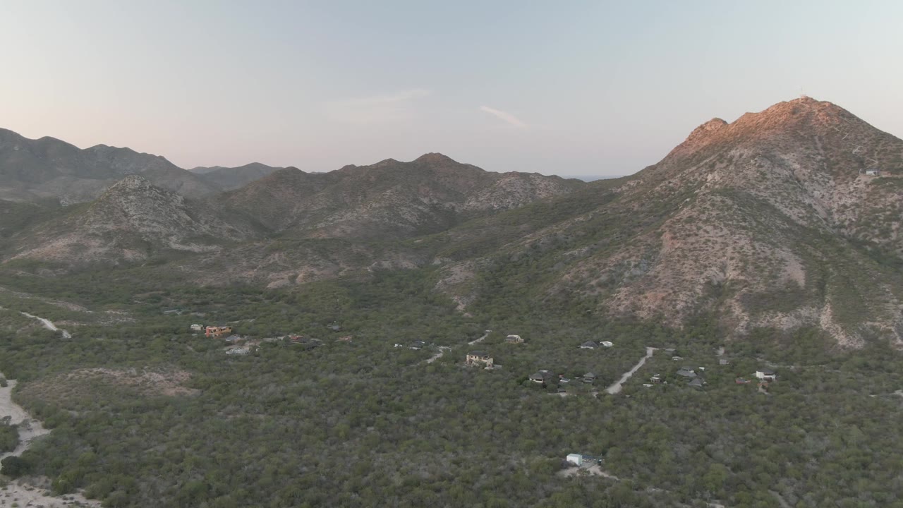 Mid-clip aerial over Cabo Pulmo shows the central rocky peak, surrounding ridgelines, green brush, branching dirt roads, and scattered structures in the valley, with no visible people.