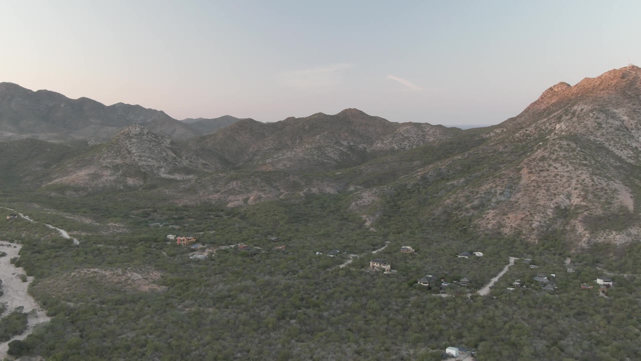 Wide aerial frame over Cabo Pulmo, Baja California Sur, Mexico, with green scrubland, pale roads, scattered structures, rocky ridges, and no visible people.