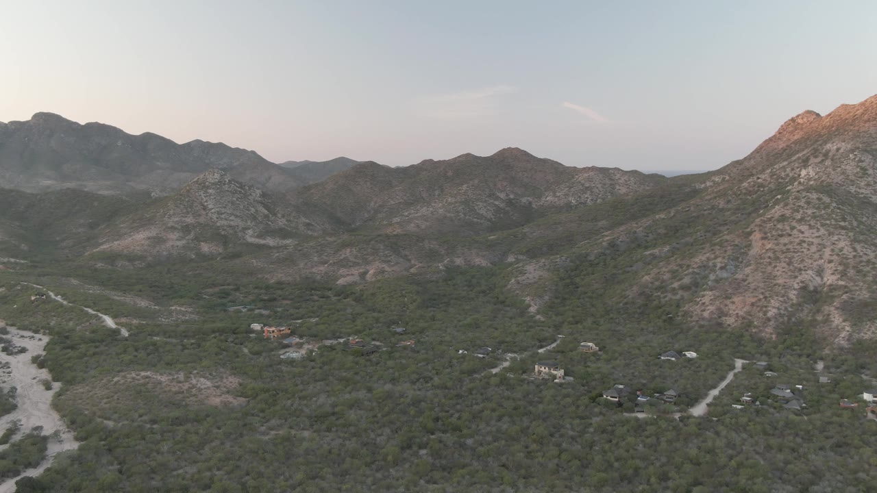 High aerial frame of the Cabo Pulmo valley with a sunlit rocky peak, layered hills, pale road cuts, scattered structures, and no visible people.