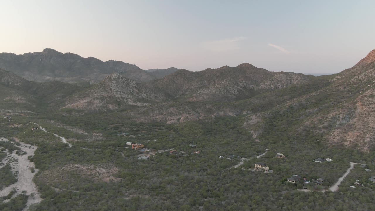 Opening aerial frame over Cabo Pulmo, Baja California Sur, with rocky mountains across the horizon, pale roads through scrub vegetation, scattered buildings below, and no visible people.