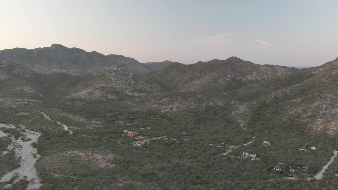 Mid-early frame in Cabo Pulmo, Baja California Sur, with the mountain ridge centered, scrubby valley terrain below, pale dirt roads curving through the landscape, and no visible people.