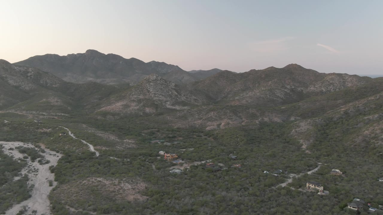 Aerial view over Cabo Pulmo, Baja California Sur, showing a pale dry wash in the lower left, green valley vegetation, rocky mountain ridges, scattered buildings, and no visible people.