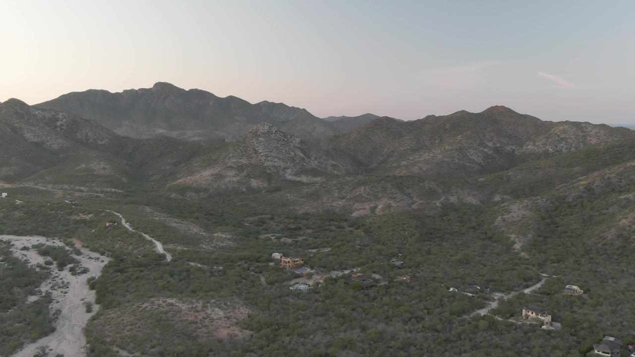 Elevated aerial frame in Cabo Pulmo, Baja California Sur, with winding dirt roads crossing green scrub, a broad rocky ridge line, scattered small structures, and no visible people.