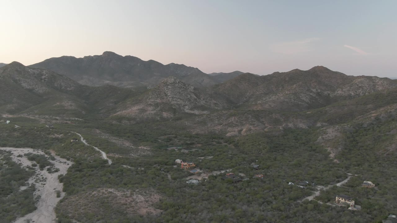Wide aerial view of Cabo Pulmo mountain terrain with green vegetation across the valley, pale dry wash areas, small buildings near the lower frame, layered rocky hills, and no visible people.