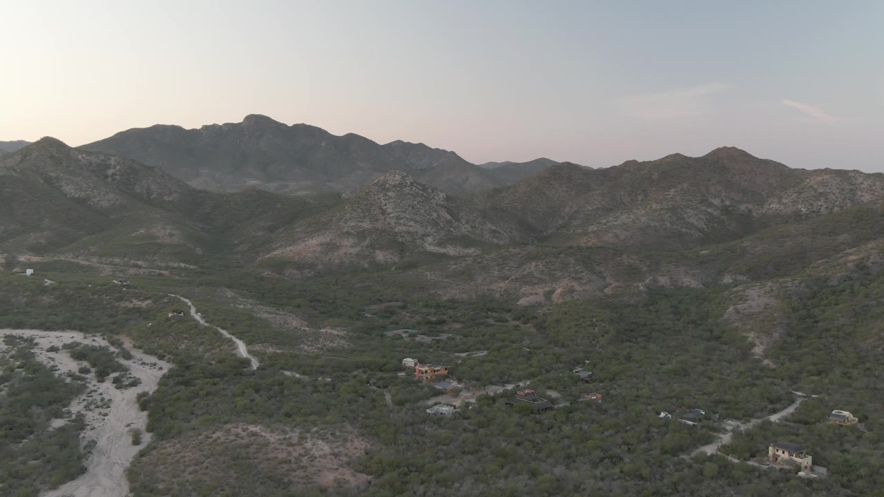 Aerial frame of dry wash, dirt road, green brush, and rugged mountains.