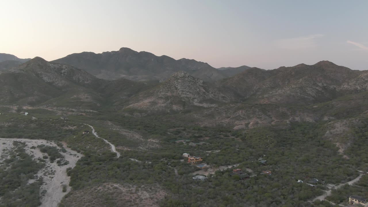 Opening aerial frame over Cabo Pulmo, Baja California Sur, showing a pale dry riverbed through scrub vegetation with rocky mountains under a hazy sky and no visible people.