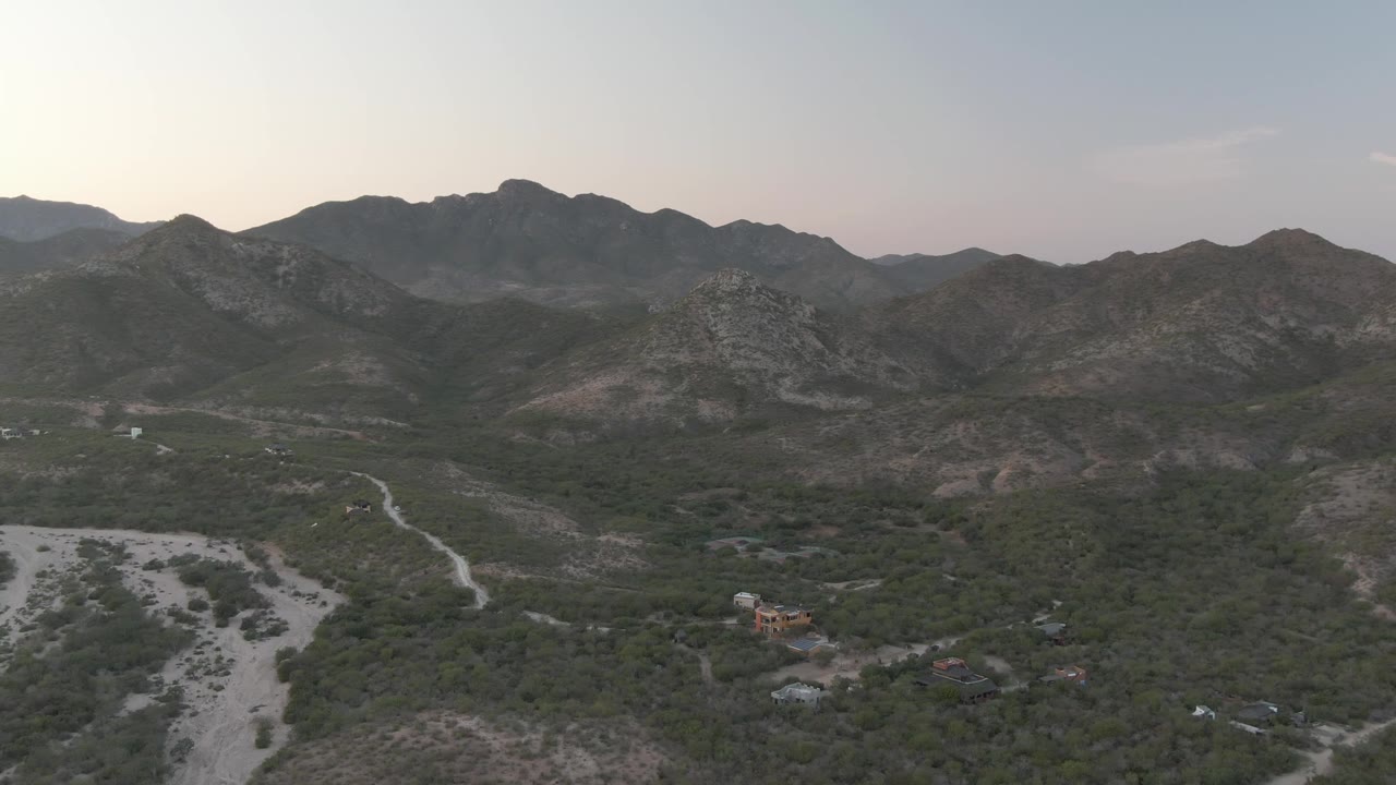 Third frame over Cabo Pulmo, Baja California Sur, showing rocky hills spanning the background, a pale river channel below, muted green vegetation, and no visible people.