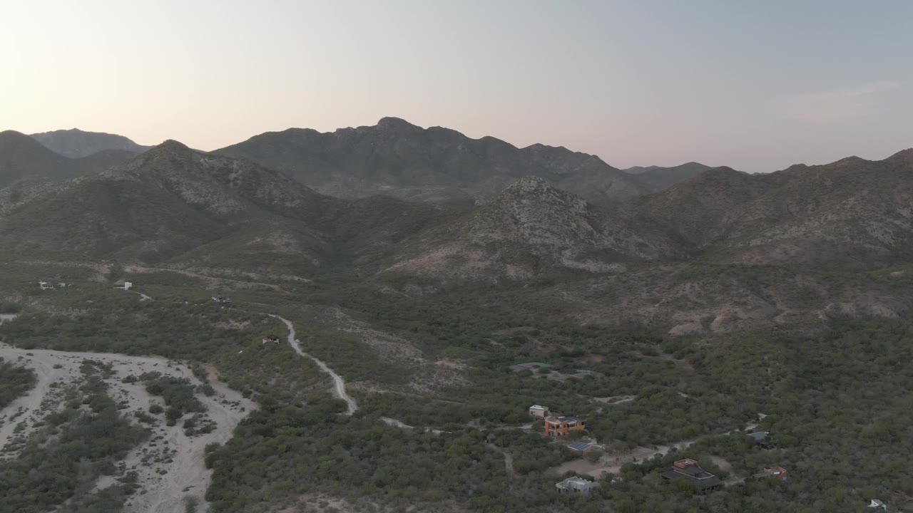 Aerial view over Cabo Pulmo, Baja California Sur, showing a pale dry riverbed, scrub vegetation, distant rocky mountains, low dusk light, and no visible people.