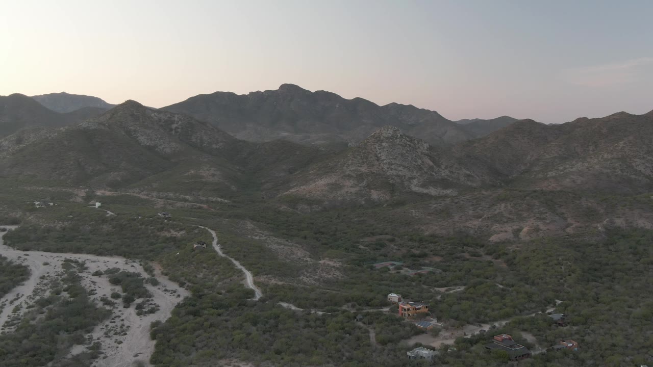 Wide drone view of scrubland, dirt paths, and rocky mountains beyond a dry wash.