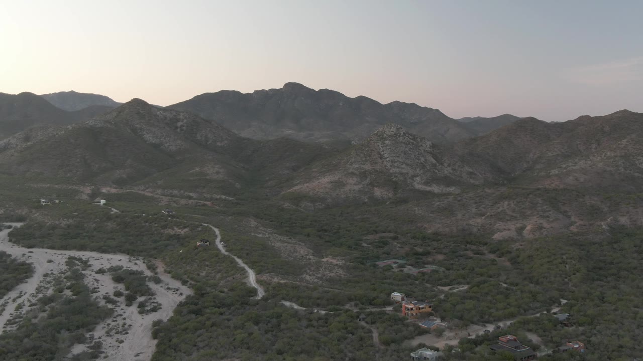 Drone-height view over Cabo Pulmo terrain with a pale dry riverbed in the lower valley, green scrub, scattered dirt tracks, layered mountain ridges, and no visible people.