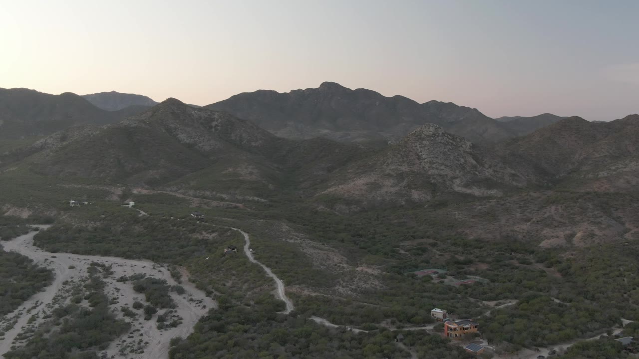 Wide aerial frame over Cabo Pulmo, Baja California Sur, showing a pale dry riverbed, curving dirt road, scrub vegetation, rocky ridges, small structures near the lower edge, and no visible people.
