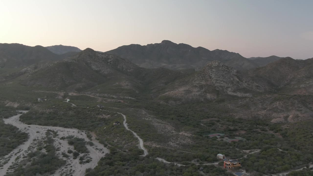 Aerial view in Cabo Pulmo, Baja California Sur, with the dry riverbed more centered below, a narrow road running through green scrub, layered rocky mountains beyond, and no visible people.