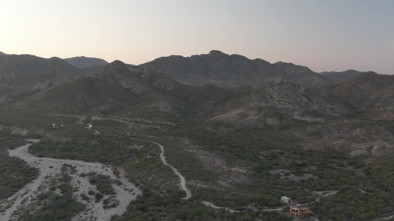 Cabo Pulmo aerial frame showing the pale riverbed branching through the lower valley, a winding dirt road on the right side, dark scrub cover, rocky mountain slopes, and no visible people.