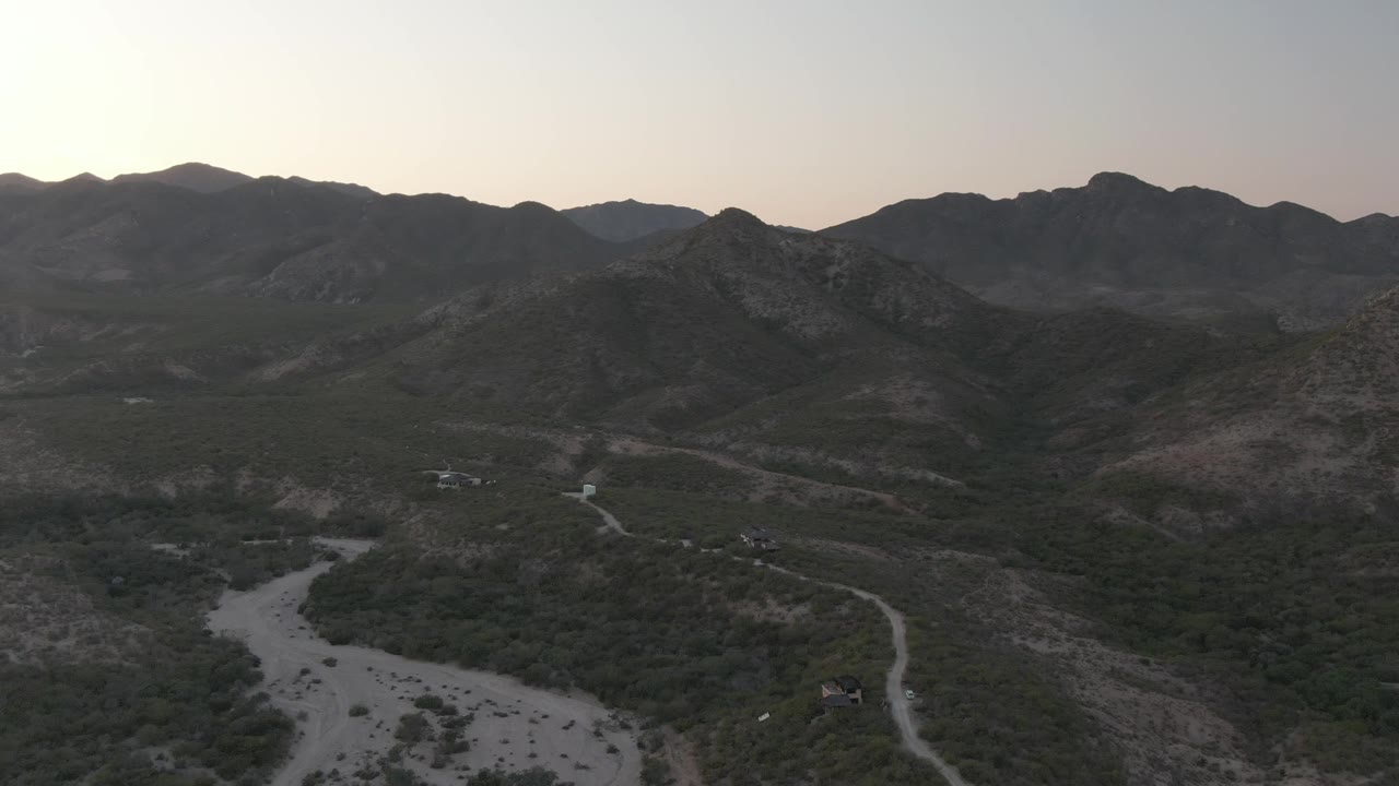 Aerial dusk view over Cabo Pulmo, Baja California Sur, with a pale dry riverbed in the lower frame, winding dirt roads through scrub, distant mountain ridges, and no visible people.