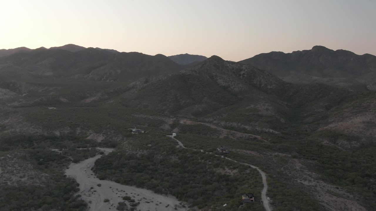 Aerial dusk view of a mountain valley with a dry wash, dirt tracks, scrub, and small structures.