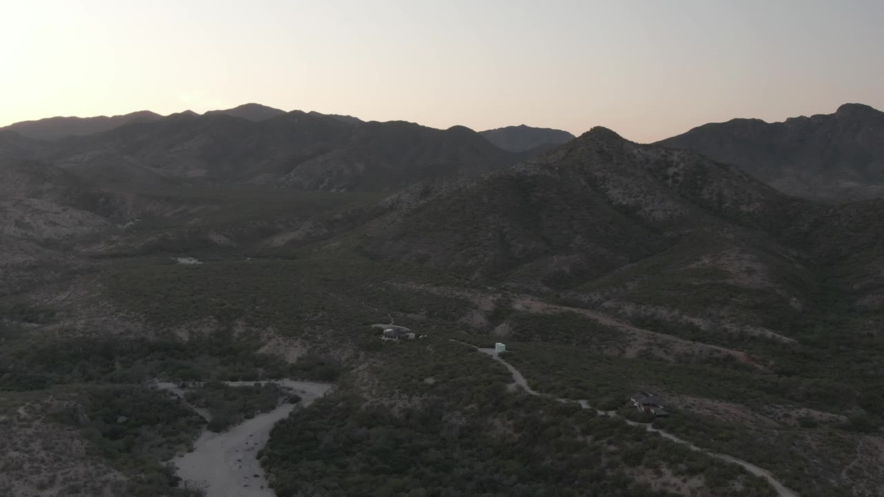 Aerial view over Cabo Pulmo, Baja California Sur, with dark mountain ridges, pale dirt roads, sparse vegetation, and no visible people at the start of the clip.