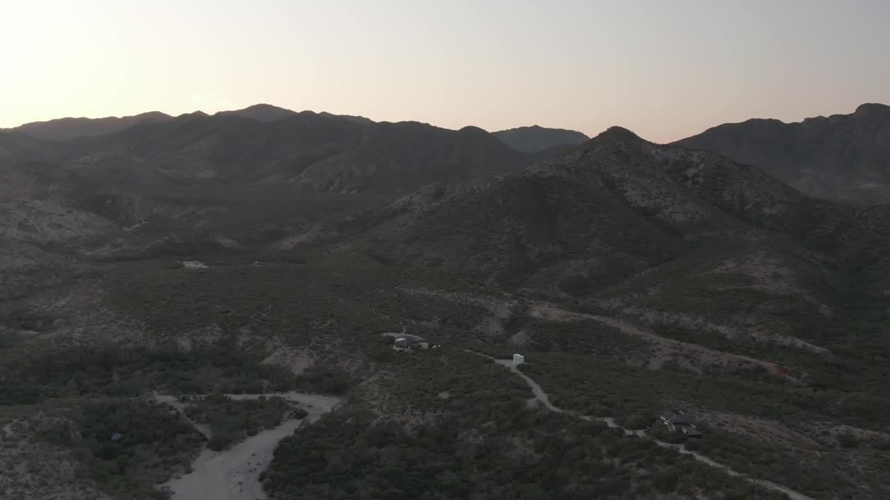 The aerial composition holds on Cabo Pulmo's winding dirt roads, scattered structures, dark hillside vegetation, distant ridgelines, and no visible people.
