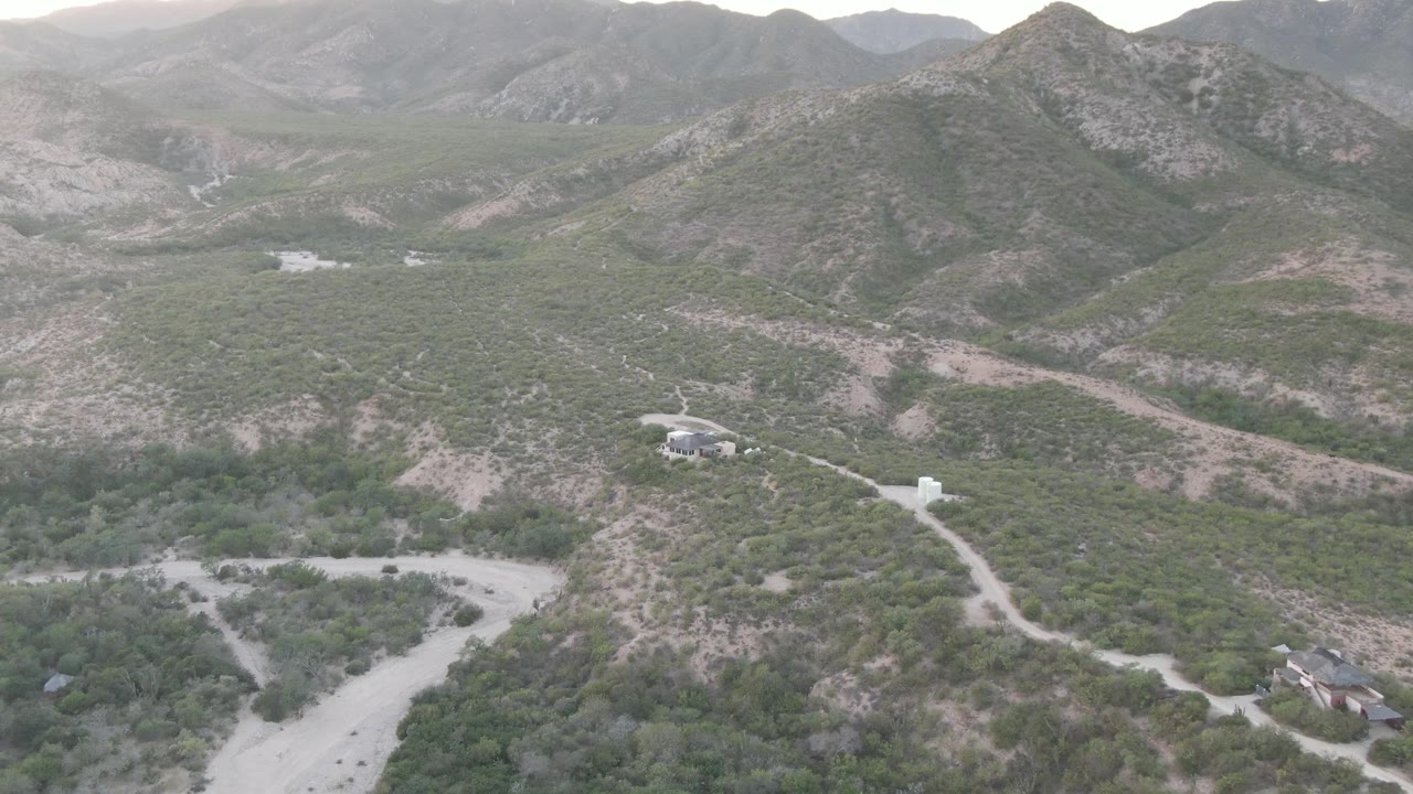 Aerial frame over Cabo Pulmo, Baja California Sur, showing pale dirt roads, low green scrub, dry hills, a few distant buildings, and no visible people.