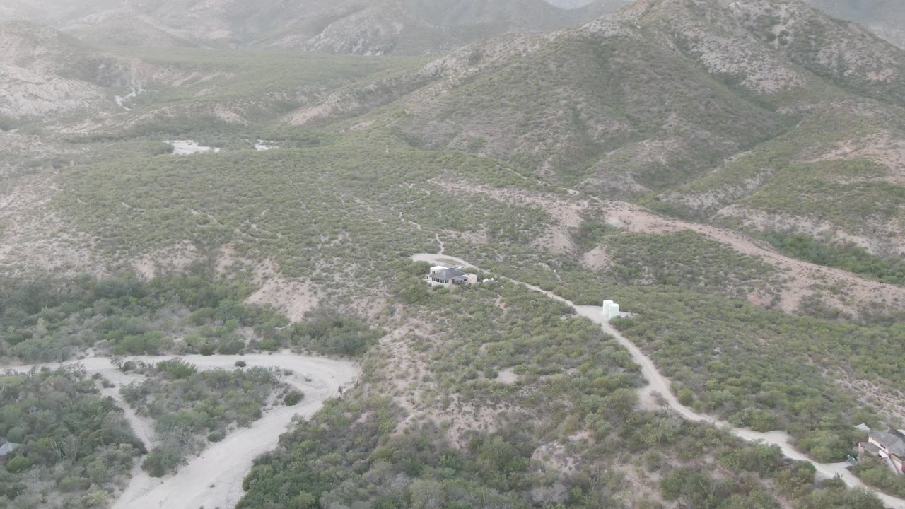 Cabo Pulmo aerial frame showing a pale dirt road rising toward scrub-covered hills, small buildings near the lower right, hazy mountain slopes, and no visible people.