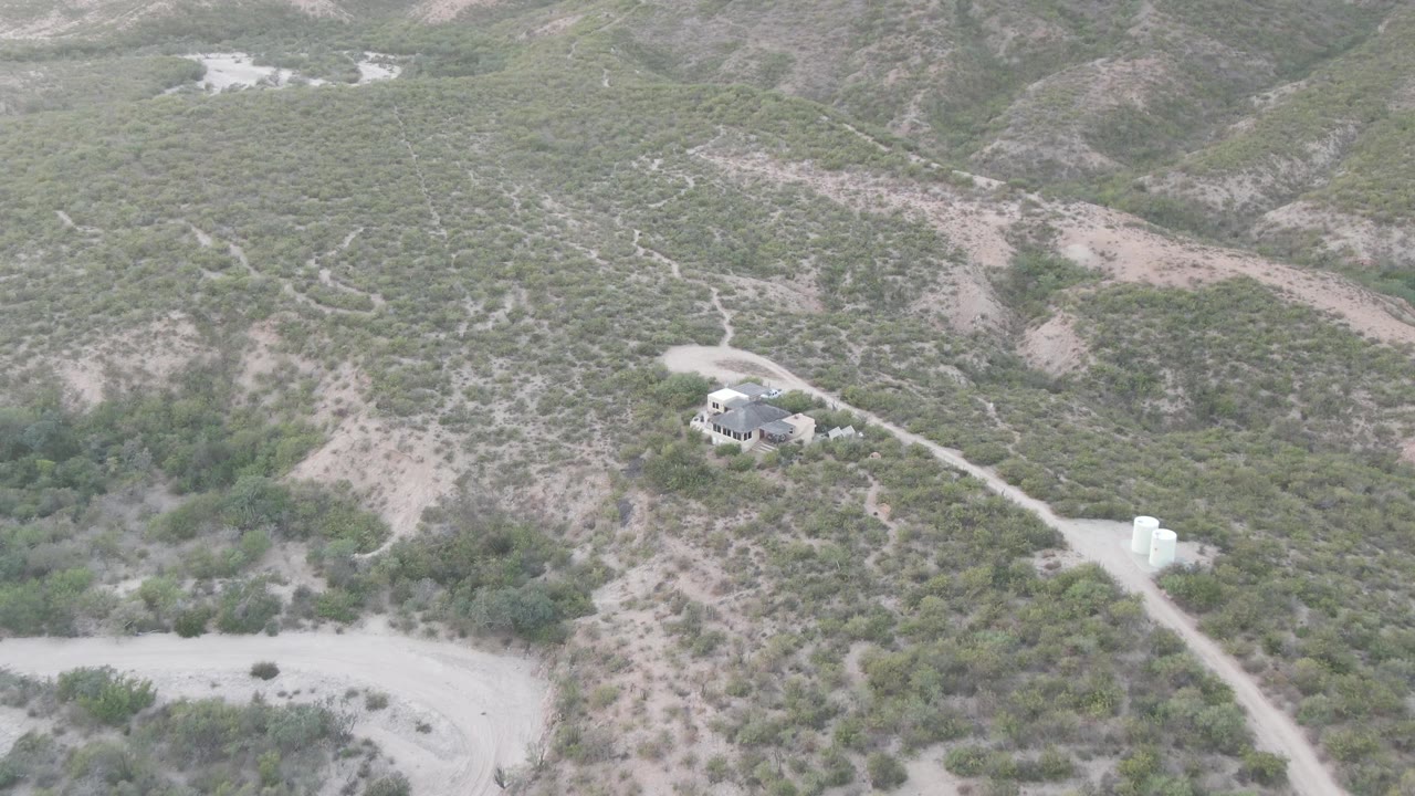 High aerial view in Cabo Pulmo, Baja California Sur showing scrub-covered arid hills, a small house near a dirt road, pale soil, and no visible people.