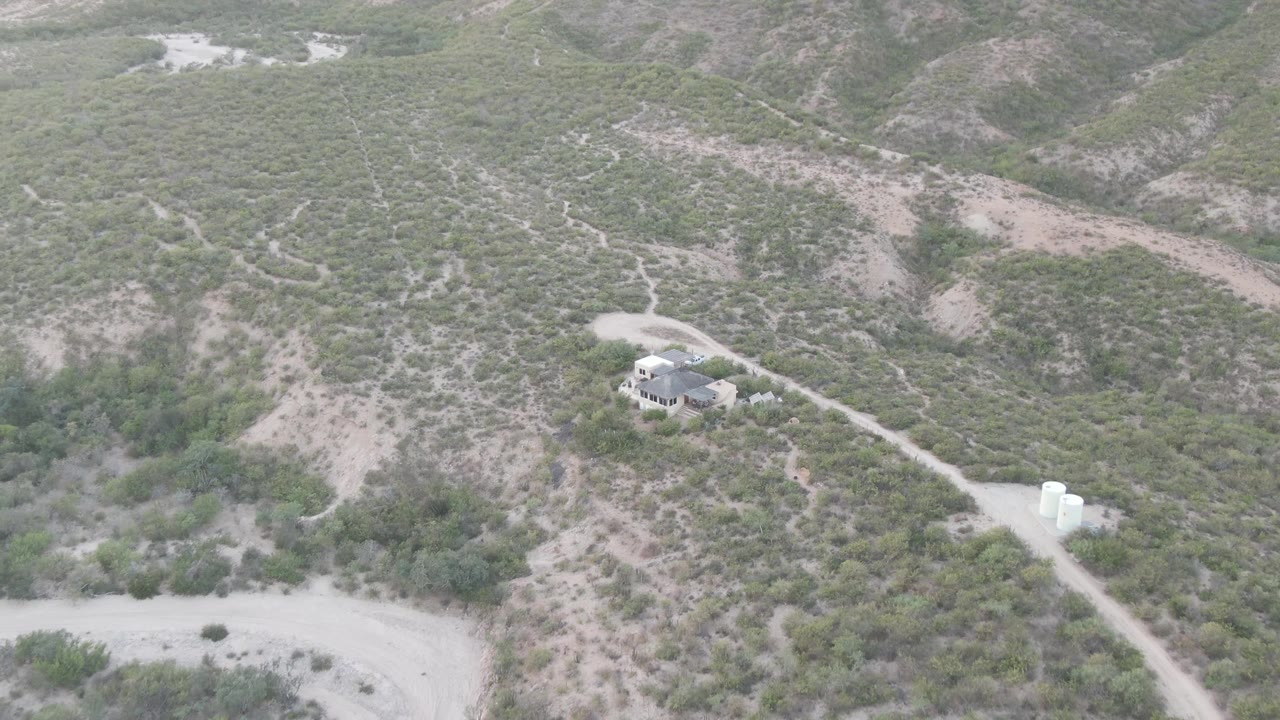 Drone view of a small house, road, and white water tanks in dry hills.