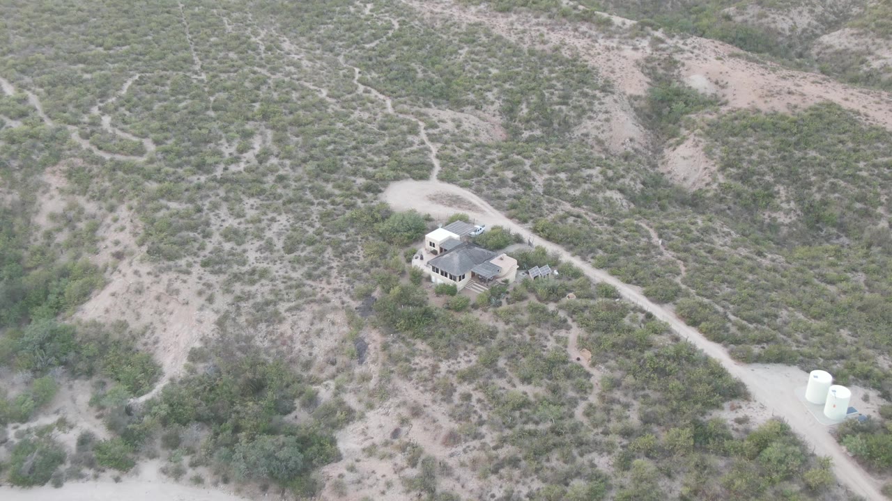 Aerial frame over Cabo Pulmo showing a small light-colored house below scrubby desert hills, a winding dirt road, white tanks at the lower right, and no visible people.
