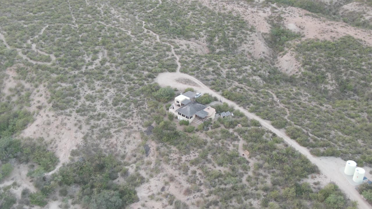 Aerial view in Cabo Pulmo with the house slightly lower in frame, the pale dirt road crossing toward white tanks, scrub vegetation across the slope, and no visible people.