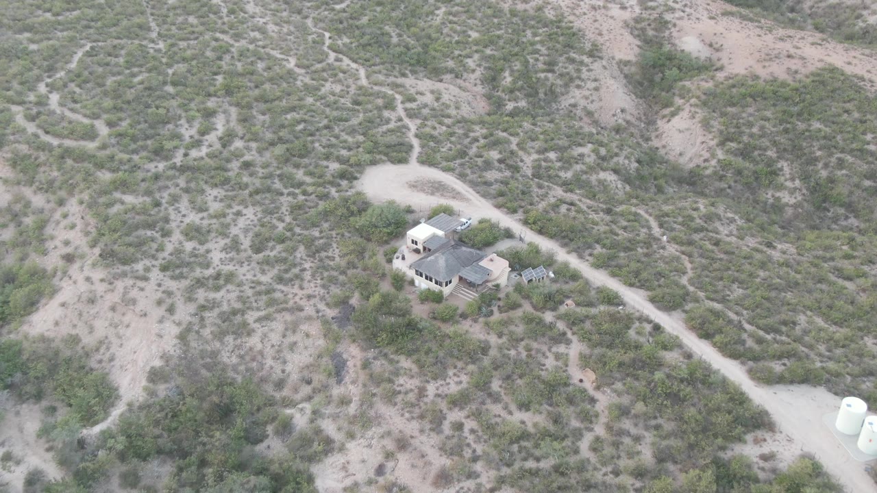 Cabo Pulmo aerial frame showing the house near the lower left, a looping dirt road above it, dry brush patterns across the hillside, white tanks at the right edge, and no visible people.
